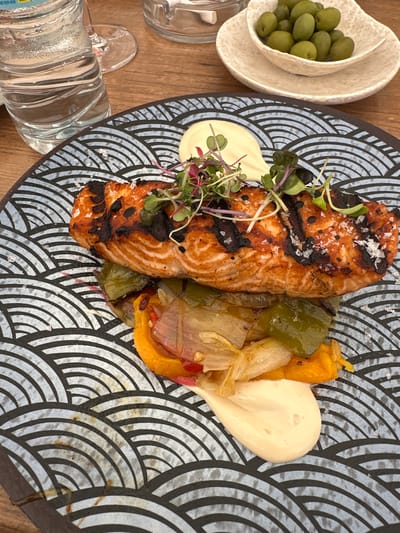 A top-down shot shows a plate of food, a bowl of olives, and a water bottle on a wooden table. The plate features a piece of grilled salmon, roasted peppers (orange, green, and red), a dollop of white sauce, and a garnish of microgreens. The salmon has grill marks and is topped with flakes of salt. The bowl of olives is partially visible in the upper left corner. The water bottle is in the lower left corner of the image. The plate is decorated with a wavy black and white pattern. The background and specific location in Sevilla, Spain are not discernible from the image.