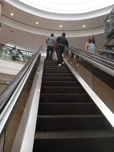 The image captures the interior of a shopping mall in Johannesburg, South Africa. The focus is on an upward-moving escalator, with three individuals visible on it. Two men are ascending: one wearing a gray t-shirt and the other wearing a black t-shirt, black pants, and white shoes. A woman in a light blue t-shirt stands at the top of the escalator. The escalator has stainless steel handrails. The background shows a multi-level structure with a curved ceiling, recessed lighting, and a seating area on an upper level.