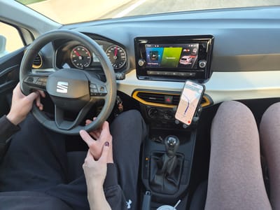 The image depicts the interior of a moving automobile, viewed from a rear passenger perspective, within the Municipal Unit of Megara, Greece.
Two occupants are partially visible. The driver's hands are positioned on the black, multi-function steering wheel, bearing a "SEAT" logo. The driver wears dark long-sleeved clothing and dark sweatpants. The driver's left hand is interlaced with the right hand of the front passenger. The passenger's legs are visible, covered in light brown leggings. The passenger's right hand displays dark red nail polish.
The vehicle's dashboard features a two-tone design of black and off-white/beige, with a distinctive yellow trim surrounding the central air vent on the passenger side. The instrument cluster shows an analog speedometer indicating approximately 50 km/h, and a digital display beneath it reads "08:00". The central infotainment touchscreen displays the time "17:45", music information including "Never Thought I'd..." and Greek text "Όχι αγαπημένα", and a graphic for "Front Assist". A smartphone is secured in a dashboard mount, displaying a navigation application (Google Maps) with a current speed of 42 km/h, a route to "Chaïdari", and an estimated arrival time of "17:39". The vehicle is equipped with a manual 5-speed gear shift. The center console contains keys on a keyring, a white cup with a straw, and an illuminated red hazard warning light button.
Through the windshield, a clear road, consistent with a highway, is visible under bright daylight conditions.