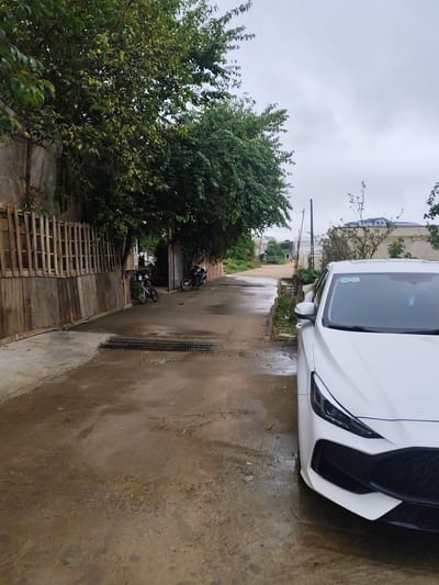 This image depicts a wet, paved alleyway in Lam Vien, Da Lat, Vietnam. A white car is parked on the right side of the frame, partially obstructing the view of the alley. The alley is lined with wooden fences and lush green trees on the left, and buildings and more vegetation on the right. Two motorcycles are parked further down the alley on the left. The sky is overcast, suggesting a cloudy or rainy day. The ground is wet and reflective, indicating recent rain. The overall scene suggests a residential or semi-commercial area within the city.