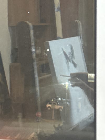 The image depicts a reflection in a window, showing an indoor scene with an artist painting on an easel. The artist's hands are visible, holding a paintbrush and a palette. The painting on the easel appears to be a figure with wings. In the background, there are shelves with various objects, and a small white truck is visible on a street through the window. A red traffic sign is also present on the street. The scene suggests an art studio or a room with artistic materials. The image does not provide specific details about the city of Armavir, Russia, as the location is not visually identifiable.