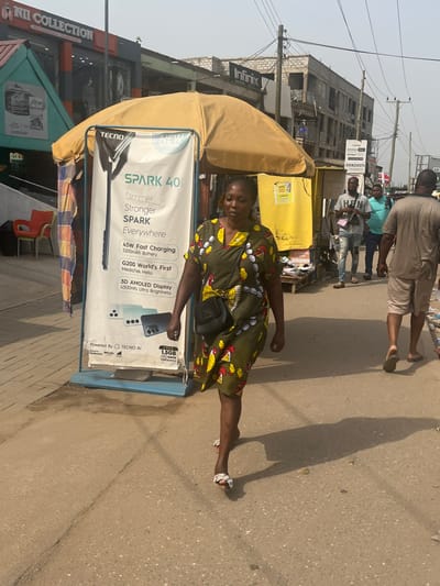 The image presents a daytime urban street scene in Ablekuma, Ghana. A woman in a dark olive-green knee-length dress with a yellow, red, and white pattern, carrying a black cross-body bag, walks towards the left foreground on a paved surface, casting a strong shadow to her right. She wears light-colored open-toed sandals. Immediately to her left is a blue-based portable advertising stand with a beige umbrella canopy, prominently displaying a "TECNO SPARK 40" advertisement with product details and a "FREE 1.5GB DATA" offer. Behind the advertising stand, to the far left, a shop labeled "NII COLLECTION" is visible with clothing displays and an orange plastic chair outside. To the right of the foreground woman, further down the street, several men are present; one in a grey t-shirt and ripped jeans with a pink cross-body bag, another in a light blue t-shirt, and a third, seen from the back, in a beige t-shirt and light shorts, wearing flip-flops. A yellow vendor stall with an "MTN" sign is partially visible behind the advertising stand. The background includes multi-story buildings, some featuring exposed concrete blocks, an "Infinix" sign on one structure, and numerous overhead utility lines and poles against a bright, hazy sky. The overall environment suggests a commercial area.