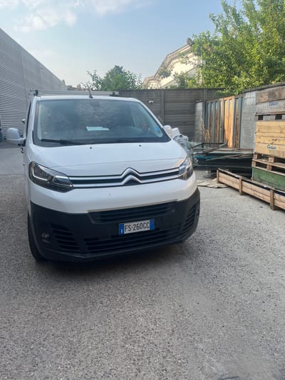 A white Citroen Jumper van with license plate FS 260CC is parked in an outdoor industrial area. The van is facing forward, with its headlights on. The background includes a corrugated metal building on the left, a wooden fence, some trees, and stacked wooden pallets on the right. The ground is paved with asphalt. The scene is under a partly cloudy sky. The location is identified as Rozzano, Italy.