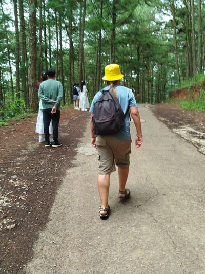 The image captures a pathway winding through a dense forest, characterized by tall pine trees and a light-colored, gravelly ground. In the foreground, a person wearing a yellow bucket hat, a blue t-shirt, and khaki shorts walks away from the camera, carrying a dark backpack. Their legs are visible, wearing sandals. To the left of the pathway, several individuals are gathered. One person in a green long-sleeved shirt and black pants stands with their back to the camera, alongside another person wearing a white dress and a third in a dark blue top and skirt. The overall setting suggests an outdoor recreational area within the city of Cam Ly, Da Lat, Vietnam, possibly a park or nature trail, given the presence of both tourists and what appear to be locals in traditional attire.
