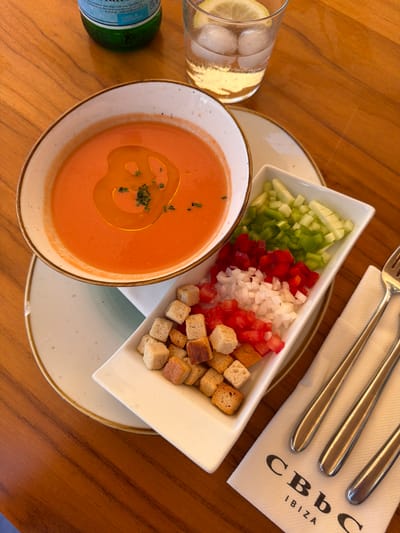 A close-up, top-down shot captures a Spanish gazpacho soup served in a rustic bowl, drizzled with olive oil and garnished with finely chopped herbs. Alongside the soup, a rectangular white dish holds a colorful medley of finely diced vegetables—cucumber, green bell pepper, red bell pepper, and white onion—as well as a pile of croutons. A glass of water with ice and a lemon slice sits to the right of the soup, and a bottle of San Pellegrino water is partially visible in the background. A white napkin with the "CBbC IBIZA" logo is placed below the vegetable dish, adorned with three silver forks. The objects are arranged on a warm, polished wooden table, suggestive of a restaurant setting in Sant Josep de sa Talaia, Spain, likely at CBbC, a beach club in Ibiza.