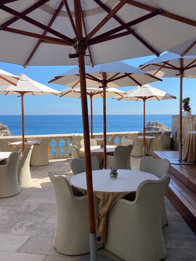This is an outdoor seating area on a terrace overlooking the Adriatic Sea in Dubrovnik, Croatia. The scene is set on a sunny day with a clear blue sky. Several white umbrellas with wooden poles provide shade over tables and chairs. The tables are round and covered with light-colored tablecloths. The chairs are woven, with a light beige or cream color.
In the background, the sea is a deep blue, and a rocky island is visible. A balustrade runs along the edge of the terrace, separating it from the sea. The terrace floor is paved with large, light-colored stone tiles. The scene appears to be a restaurant or cafe setting, designed for al fresco dining with a scenic view. There are no people visible in the image. The overall atmosphere is serene and tranquil, highlighting the beautiful coastal location.