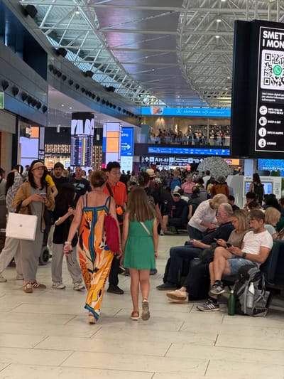 The image shows the interior of an airport, likely at Fiumicino Airport near Rome, Italy. Several people are standing and sitting, seemingly waiting for their flights or baggage. There are information boards displaying flight details and other announcements. Several shops and eateries are visible, and there is signage indicating directions to gates A31-83. A cafe called "ALEMAGNA caffè pasticceria" is seen. Overhanging electronic signs display QR codes and airport information. The architectural structure of the airport ceiling is visible.