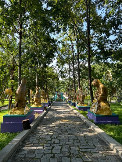 This is a low-angle, eye-level outdoor shot of a stone pathway leading through a park in Thuận Nam, Vietnam. The pathway is flanked by lush green grass and tall trees with dense foliage. Along the right side of the pathway, a series of golden statues of animals are displayed on purple and green pedestals. These statues appear to be part of the Chinese zodiac, with figures such as a monkey, a cat, and a bull visible. The statues are adorned with decorative white cloud motifs. The pathway is paved with irregular grey cobblestones, and sunlight casts dappled shadows across it. In the distance, the pathway curves and leads towards what appears to be a decorated archway or structure. The sky is visible above the trees, appearing clear and blue.