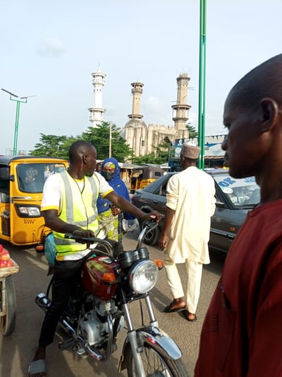 In Arawa, Nigeria, a man wearing a yellow and green reflective vest is on a motorcycle, looking to his right. He is wearing black pants and grey sandals. Behind him, a woman in a blue headscarf and patterned dress is partially visible. To the left of the motorcyclist is a yellow tricycle taxi. In the background, a mosque with multiple minarets stands under a bright sky with scattered clouds. Several other vehicles, including a dark-colored car and other tricycles, are present on the road. A man in a light-colored robe and cap stands near the car, facing away from the camera. The scene appears to be a busy street with traffic.