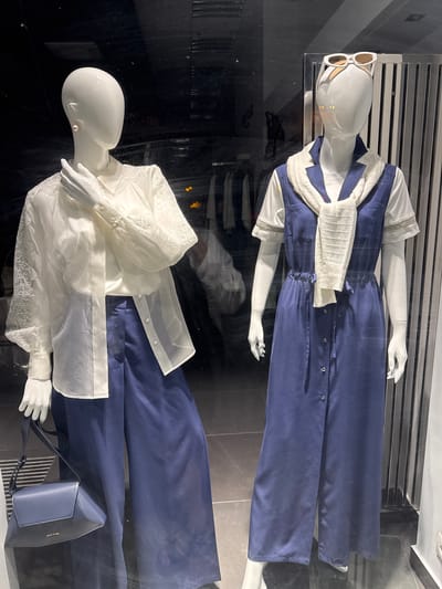 This image displays two mannequins in a retail setting, likely a clothing store window. The mannequins are dressed in a dark blue, full-length ensemble that appears to be a jumpsuit or dress with wide legs. The top part of the garment is partially obscured by a white, textured sweater or scarf draped around the shoulders.

The top mannequin wears large, white-framed sunglasses. The lower mannequin is adorned with a sheer, white blouse featuring lace details on the sleeves and yoke. It is also accessorized with a dark blue clutch purse. The background consists of reflective surfaces, suggesting a polished floor and potentially a display structure. The lighting is artificial, creating a bright, shop-like ambiance. There is no indication of the time of day or weather conditions. The overall scene evokes a sense of curated fashion display. The clutch has "DIESEL" and "ARMY" in gold lettering.