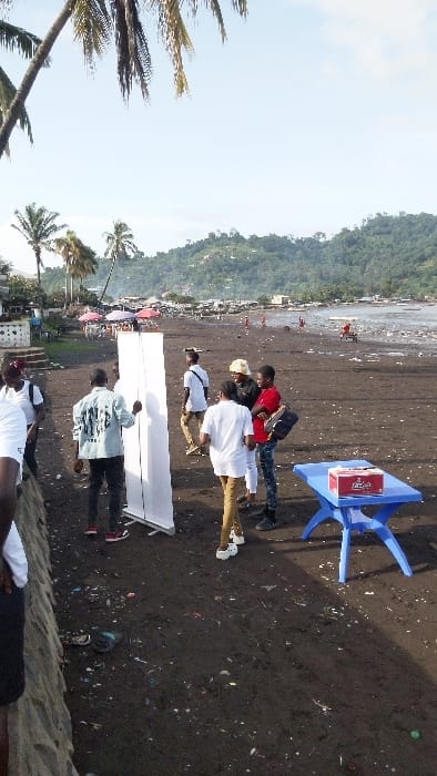 The image depicts a scene on a beach in Limbe, Cameroon (latitude: 4.004, longitude: 9.211). Several people, mostly men, are standing around a large, white, freestanding, retractable banner and a blue, plastic table. Some people are talking to each other, while others are looking at the banner. The beach is covered in dark sand and some debris. There are also many palm trees and other vegetation in the background. The image is taken from the perspective of a person standing close to the banner. The scene is framed by a palm tree on the left and a green hillside in the distance.