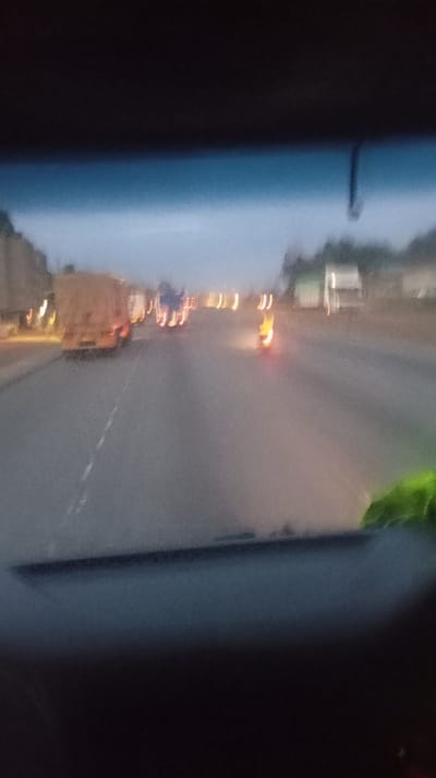 A blurry, out-of-focus view from inside a vehicle looking out onto a road. Several vehicles, including what appear to be trucks, are visible on the road ahead, along with blurred lights. The sky is overcast or dusk is approaching. A portion of the vehicle's dashboard and windshield are visible in the foreground, adding to the obscured perspective. The overall impression is of motion and unclear visibility.