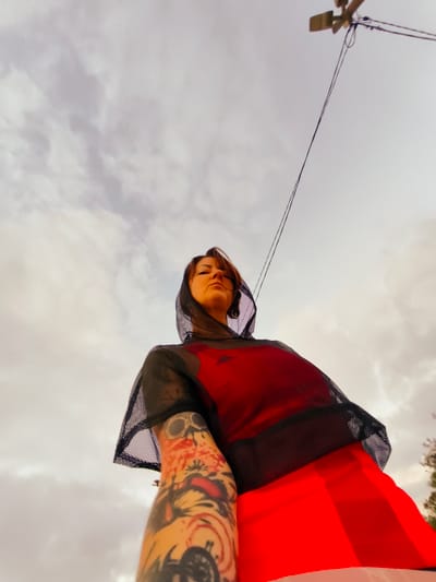 The image depicts a low-angle shot of a woman wearing a red garment and a black mesh hooded shirt. She has tattoos on her left arm and a nose ring. The background consists of a cloudy sky with visible power lines and a utility pole. Trees are also visible in the distance on the right side of the frame. The scene appears to be outdoors in a non-urban area.
