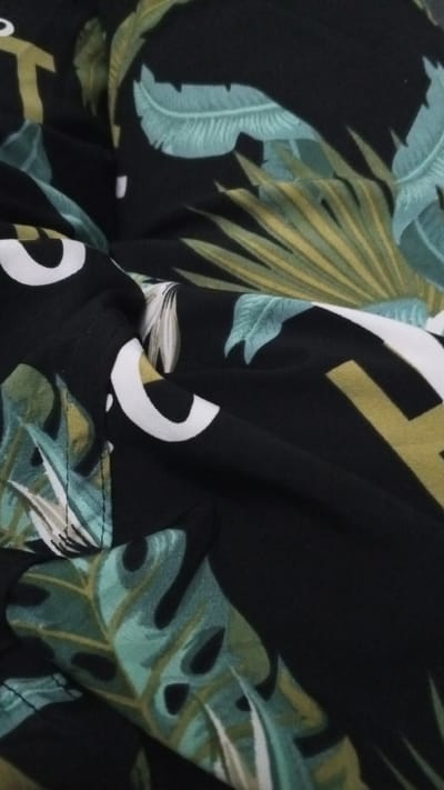 The image displays a close-up, abstract view of a black fabric with a tropical leaf print. The leaves are rendered in shades of green, teal, and yellow, with some white accents. The fabric is wrinkled and folded, obscuring any clear sense of a larger object or scene. There are no people, identifiable locations within Dushanbe, Tajikistan, or any discernible actions taking place. The focus is entirely on the texture and pattern of the textile.