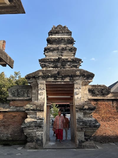 Two men are walking through an ornate stone gateway. The man in the foreground is wearing a red and white patterned shirt and a red sarong. He is walking towards the camera. The man behind him is wearing a brown shirt and white pants, and is walking in the same direction as the foreground man. The gateway is constructed of brick and stone, with intricate carvings. The sky is blue and clear. The scene appears to be the entrance to a temple or mosque in Yogyakarta, Indonesia.