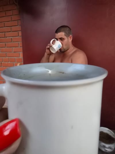 In Nova Kakhovka, Ukraine, a man with short dark hair and a beard is standing shirtless, holding a white mug to his lips and drinking from it. He is positioned in front of a dark red wall. To the left of the man, a brick wall is visible. In the foreground, a large, white mug with a thick rim is prominently displayed, partially obscuring the man and the background. The mug appears to contain a clear liquid. A person's hand, with a bright red fingernail, is holding the large white mug.