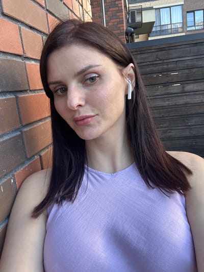 A young woman with long, dark hair is lying on a brick surface, her head tilted. She is wearing a light purple, sleeveless top. She has earbuds in her ears. The background shows a wooden fence and a modern building with windows. The lighting suggests it is daytime. The scene appears to be outdoors, possibly in an urban or suburban setting. Her expression is neutral.