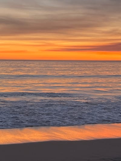 The image depicts a serene beach scene at sunset, with the sky a vibrant canvas of orange and purple hues. The ocean stretches out to the horizon, reflecting the warm colors of the sky. Gentle waves lap at the shore, creating a foamy white edge against the dark sand. This photograph was taken at Praia de Riazor, a popular urban beach located in the city of A Coruña, Spain. No people or specific objects are visible in the frame, emphasizing the natural beauty of the coastal landscape.