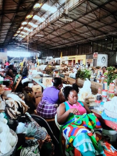 An interior view of a bustling market in Ihumwa, Tanzania, is presented, characterized by a high, industrial-style metal truss roof with several translucent panels serving as skylights. The market floor is densely packed with numerous vendor stalls, displaying a wide variety of goods. In the immediate foreground to the right, a dark-skinned woman with dreadlocks wears a pink top and a vibrant multi-colored wrap/skirt (featuring green, blue, red, yellow, and white patterns). She is seated in a white plastic chair, looking downwards, with her right hand visible. Behind her, another dark-skinned woman in a purple striped top and a purple and white plaid wrap is seated on a low wooden stool, facing away from the camera.
The stalls throughout the market feature large quantities of produce, including green leafy vegetables, root vegetables, and other items displayed in piles, baskets, and on tables. Various containers, such as white plastic buckets, clear plastic bottles containing liquids or spices, and cardboard boxes, are visible on and around the stalls. The market appears crowded with indistinct figures of people engaged in buying and selling activities. In the distant background, signs and posters are affixed to walls, including a prominent black and white image on a white surface.