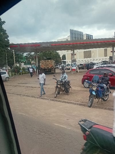 The image depicts the forecourt area of a TotalEnergies gas station located in Nanyuki, Kenya, under an overcast sky. The view is from a low angle, seemingly from within a vehicle, with the top of a vehicle's dashboard or interior panel partially visible in the bottom right foreground.

In the immediate foreground, a cracked asphalt road surface with a metal drainage grate is visible. To the right, a dark motorcycle with a red seat and taillight is partially visible.

In the midground, three individuals and two motorcycles are prominent. A man wearing a light-colored short-sleeved shirt and blue jeans walks from left to right on a paved surface. To his right, another man, dressed in a plaid shirt and dark pants, sits astride a brown motorcycle parked on the paved area. Further to the right, a third man, wearing a dark jacket and blue jeans, is seated on a blue motorcycle, also parked on the paved surface adjacent to a red sedan.

The background features the grey canopy of the TotalEnergies station, with the "TotalEnergies NANYUKI" logo clearly visible in red and white lettering. Beneath the canopy, several gas pumps are discernible, and various vehicles are parked or positioned for service, including a large brown dump truck near the center, multiple white cars and SUVs, and another silver car on the far left. Behind the gas station, a multi-story building with a modern, glass-paneled facade rises, and various trees and foliage are visible on the left side of the frame. The ground within the gas station forecourt appears to be a mix of paved sections and unpaved dirt or gravel.