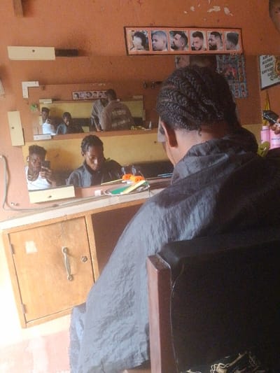 The image captures a scene inside what appears to be a barbershop or hair salon, viewed from behind a customer. The main subject, seen from the back, has intricate braided hair (cornrows or similar style) and is wearing a dark, possibly black, barber's smock or cape. They are seated in a wooden chair, facing a large mirror that reflects the activity in the room.
In the mirror's reflection, directly in front of the main subject, is another person with braided hair, likely the barber or stylist. This person is looking downwards, seemingly focused on the main subject's hair or something on the counter in front of them. To the left in the reflection, visible in a smaller segment of the mirror or a different mirror panel, a young man with dark, curly hair is taking a selfie with a smartphone held in his right hand. He is wearing a white tank top and wired earphones.
Further back in the central mirror's reflection, behind the barber/stylist, another person is visible from the back, wearing a light-colored shirt or smock. On the back of this person's attire, text is visible, which appears to read "MOLISHIDAD" or "MOLUSBID" above "POLISIDAD" or "POLISIBAD". This could be part of a uniform or a brand name.
The setting features warm, orange-brown walls, which show some signs of wear. Below the main mirror is a wooden counter with a single-door cabinet featuring a metal handle. On the counter, various items are scattered, including a round, light-colored tray, what looks like an orange hair clip or tool, and a pink bottle, possibly containing hair product, on the far right.
Above the main mirror, a framed poster displays six different male hairstyles or headshots. Below these images, faint text "POLAI??" is partially visible. To the right, on the wall, another poster is partially visible with text that includes fragments like "DE...", "BODY HAIRE...", "BABY & P...", and "SHAMPOO...", suggesting it might be an advertisement for hair care products or services.
The overall lighting suggests an indoor setting during daytime. The atmosphere appears casual and routine for a barbershop.