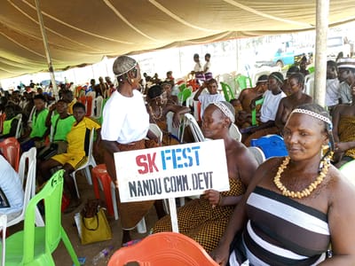 Here is a detailed description of the image:
The image shows a group of people gathered under a large tent, likely for an event or meeting. The tent is supported by poles and provides shade for the attendees. The location is Takau, Nigeria.
The people are sitting on a mix of plastic chairs, some of which are green, white, and orange. Many of them appear to be adults. Some are wearing traditional African clothing. One woman is holding a sign that reads "SK FEST NANDU COMM. DEVT". The people are casually dressed in t-shirts, dresses, and other garments. Some wear jewelry such as necklaces and headbands. The environment appears to be a public gathering. The atmosphere appears lively with some people interacting with each other.
In the background, there are vehicles parked, including a blue truck.