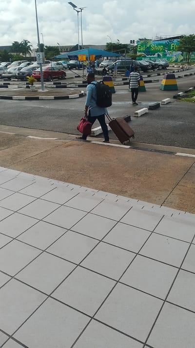 The image shows a man walking with luggage on a paved area, likely an airport or transportation hub. He is wearing a blue shirt and dark pants, with a backpack on his back and carrying a red duffel bag in his left hand. A brown wheeled suitcase is being pulled behind him by its handle. In the background, there are several parked cars, a blue canopy structure with signs, and a large billboard with green lettering that reads "Eat Shop Fly". The ground is a mix of asphalt and concrete, with white markings and curbs. There are also some decorative barriers with yellow and green stripes. The sky is overcast with scattered clouds. This scene could be at the Benin City Airport, which serves Orerokpe, Nigeria.