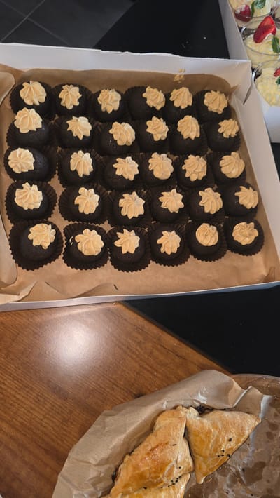 In Trikomo, Northern Cyprus, a white cardboard box contains approximately 24 chocolate truffle desserts, each individually placed in a dark brown paper liner and topped with a swirl of light brown frosting. To the right of the box, two clear plastic cups hold a creamy dessert garnished with a strawberry slice and mint leaves. In the foreground, on a wooden surface, several triangular baked pastries are arranged on brown parchment paper. The pastries appear golden brown and have a flaky crust, with visible black seeds sprinkled on top. The background is dark and out of focus.