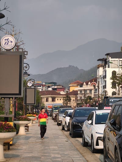 The image depicts a daytime street scene in Sa Pa, Vietnam. A paved pedestrian sidewalk, adorned with large white planters containing colorful flowering plants and wooden benches, runs along the left side. On the far left, a tall grey sign structure features a prominent white circular logo with blue abstract shapes and a large, blank rectangular display panel below it. Decorative metallic structures resembling bare trees with lights are integrated into the signage.
A lone East Asian male, wearing a red, yellow, and green horizontally striped polo shirt, black track pants, and light-colored flip-flops, walks from left to right on the sidewalk, looking forward. His hair is short and dark.
To the right of the sidewalk, a line of parked cars extends into the distance. Visible vehicles include a black SUV, a white sedan, and parts of other vehicles, including one with a "TAXI" sign on its roof.
Beyond the parked cars, multi-story buildings line the street. Several appear to be hotels, identified by signs such as "LEGACY HOTEL" and "TRAVEL." These buildings exhibit varied architectural styles, including white facades with black balconies, arched windows, and tiled roofs. Ornate black streetlights with decorative scrolls and globes illuminate the street.
In the background, large, hazy mountains rise against a light grey sky, characteristic of the mountainous terrain surrounding Sa Pa. The overall lighting suggests either an overcast day or late afternoon.