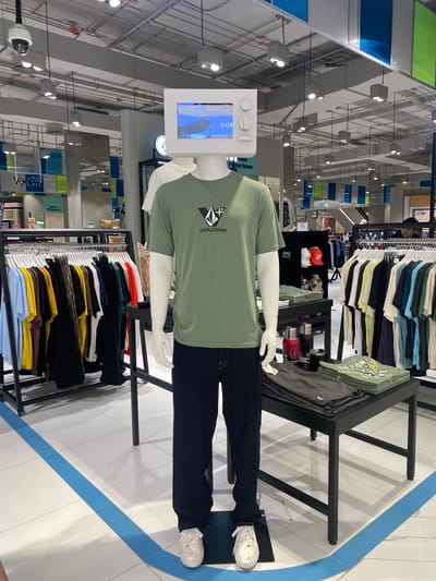 The image shows a retail store interior in Wichit, Thailand. The primary subject is a mannequin dressed in a green t-shirt and dark pants, displayed in a clothing store. The mannequin has a tablet mounted on its head, displaying a blue and white graphic. A rack of folded t-shirts in various colors is visible behind the mannequin, and another rack of t-shirts is displayed on a shelf above. A metal shelving unit with water bottles and hats is also present. The store has a modern design with white tiled floors and white ceilings with exposed ductwork and lighting fixtures. Blue and green colored panels are visible on the walls in the background. The lighting suggests it is daytime. There is no visible text on the mannequin or its clothing, but there is text on signs in the background, including "Fitting Room" and "Roxy".