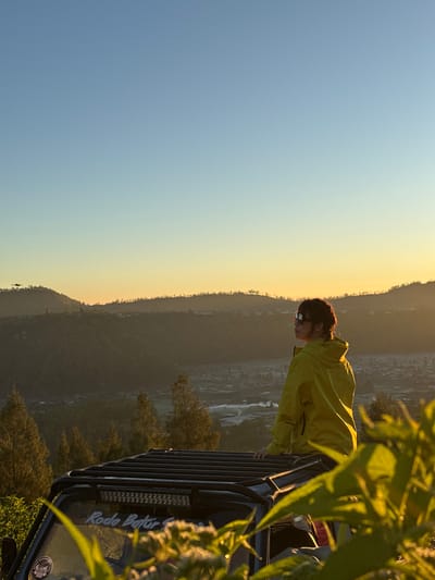 A young woman with braided hair, wearing a bright yellow jacket and sunglasses, is seated on the roof of a black off-road vehicle. She is looking out at a panoramic view of a valley and mountains. The sun is low on the horizon, casting a warm, golden light over the landscape. In the distance, a small settlement is visible, nestled amongst the rolling hills. The vehicle has a roof rack with LED lights and a sticker that reads "Roda Batur Giri". The foreground is filled with lush green foliage, some of which is in focus, creating a soft blur. The sky is a gradient of pale blue and soft yellow, indicating either sunrise or sunset. The overall scene evokes a sense of adventure and tranquility.