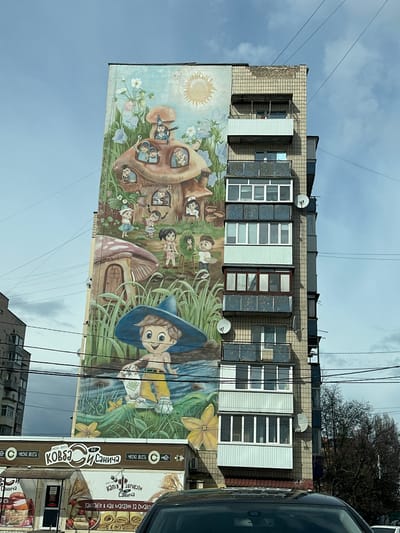 The image shows a tall building in Irpin, Ukraine, adorned with a large mural on its facade. The mural depicts a whimsical scene with children living in a mushroom house, set amidst a landscape of flowers and greenery. A boy with a blue hat is also featured prominently. Above the mural are several floors of the building with windows and satellite dishes on the balconies. Below the mural, there's a sign for "Kovbas Sanich" (Sausage Sanich), indicating a shop or business. The sky is visible with a partly cloudy appearance. A car is partially visible on the right side of the image.