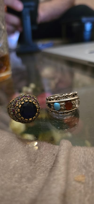 Two rings are displayed on a reflective surface that appears to be a glass tabletop. The ring on the left is ornate and gold-toned, featuring a large, deep blue stone in the center, surrounded by smaller red and clear stones. The ring on the right is silver-toned and has a more minimalist design, incorporating several smaller colored stones, including a turquoise blue one and a reddish-orange one. The background is blurred, showing hints of people and possibly a laptop, suggesting an indoor setting. The environment does not provide any specific visual cues to identify the location as Baku, Azerbaijan.
