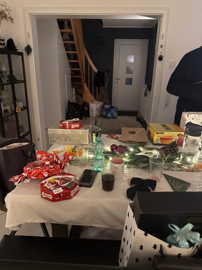A table covered with a white tablecloth displays a festive arrangement of gifts, glassware, and decorations. Presents wrapped in Christmas-themed paper, a box of One Piece merchandise, and a pinecone centerpiece with string lights are visible. The table setting includes clear glasses, some containing liquid. In the background, an open doorway reveals a staircase and a room with a closed door, giving a glimpse of the home's interior. A cabinet sits below the table, holding various decorative items.