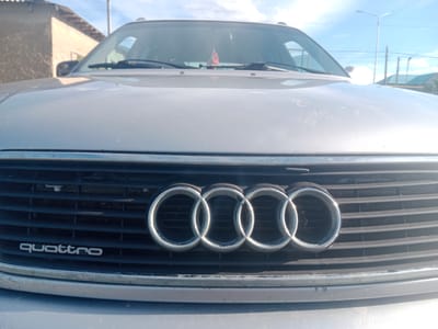 The image shows a close-up frontal view of the grill of a silver Audi car. The iconic four interlocked rings of the Audi logo are prominently displayed in the center of the grill, with the word "quattro" written in silver below it. The car is parked outdoors, with a building and power lines visible in the background under a bright blue sky with some clouds. The lighting suggests it is daytime. The specific location is Qobda, Kazakhstan.
