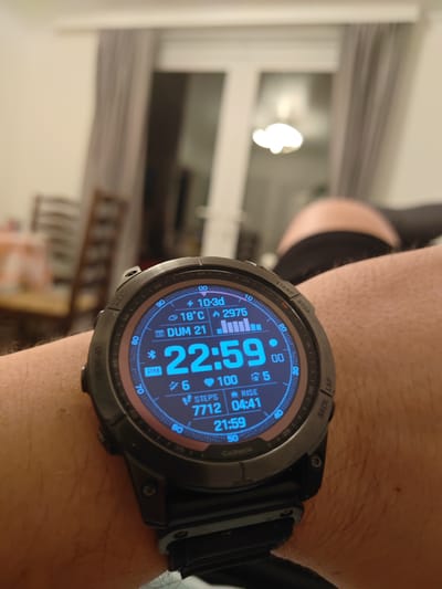 A close-up, low-angle shot captures a smartwatch on a person's wrist. The smartwatch face displays the time as 22:59, along with other data including temperature (18°C), steps (7712), and date (DUM 21). The watch is black with a blue display. The background is blurred, showing an indoor setting with curtains, a light fixture, and furniture. The person's arm is hairy and appears to be resting. There is no identifiable location in Northampton, United Kingdom.