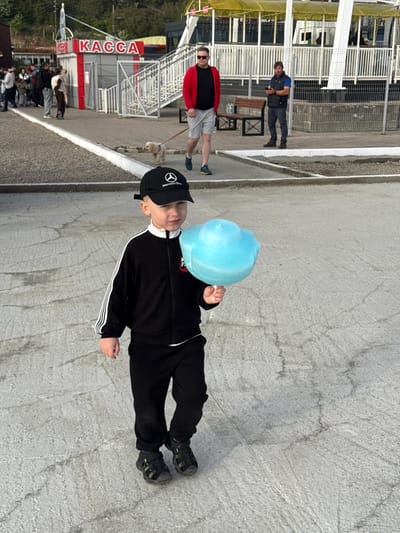 In Listvyanka, Russia, a young boy wearing a black tracksuit and a black baseball cap with a Mercedes-Benz logo walks on a paved surface, holding a large, fluffy, light blue cotton candy. Behind him, several people are visible near a ticket booth with "KACCA" written in red letters. In the background, a yellow awning covers a seating area. Two men are also in the background: one in a red jacket and grey shorts, and another in a dark vest and blue shirt. A small, light-colored dog on a leash is walking on the pavement.