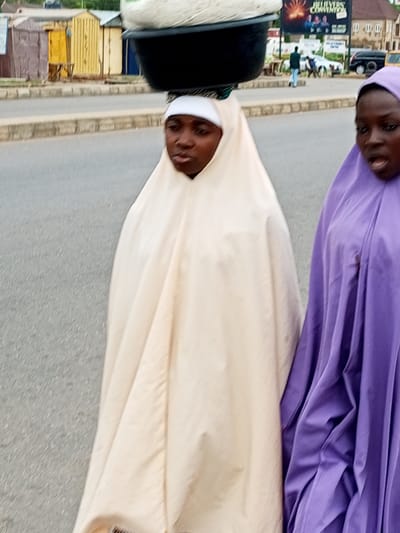 The image captures a street scene in Tumfure, Nigeria. Two women are the primary subjects. The woman on the left is wearing a cream-colored hijab and a head wrap, with a large black basin containing white material balanced on her head. She is looking forward with a neutral expression. The woman on the right is wearing a purple hijab, her mouth is open, and she appears to be speaking or calling out. They are walking on a paved road, with a sidewalk and some buildings visible in the background. The buildings appear to be small shops or stalls, painted in various colors including yellow, brown, and blue. A billboard with text and images is also visible in the background, as well as a black SUV and a yellow vehicle. The overall environment suggests an outdoor, possibly commercial area within the city.