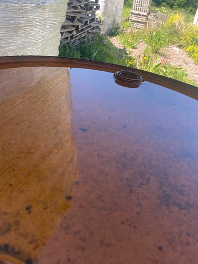 The image captures an outdoor scene in the village of Ter volevo, Russia. In the foreground, the rusted metal surface of a barrel is visible, partially filled with murky water. A circular metal fitting protrudes from the water, near the edge of the barrel. To the left, the textured side of a large, white, plastic-wrapped object, possibly a bale of material, is reflected in the water. In the background, stacks of wooden pallets and lush green grass with yellow wildflowers are visible. The overall impression is one of an industrial or agricultural outdoor storage area under a bright sky.