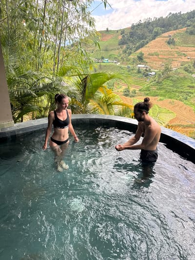 An outdoor, eye-level shot depicts a man and a woman in a dark-tiled circular hot tub or small pool, set against a backdrop of lush green foliage and terraced mountainsides in what is consistent with Mù Cang Chải, Vietnam. The woman, on the left, stands hip-deep in the water, facing right and looking down, wearing a black Nike sports bra bikini top and matching bottoms. A pink wristband is on her right wrist. The man, on the right, stands waist-deep, facing left and leaning forward, cupping water in his hands. He has dark hair tied in a top knot, a beard, and wears dark swim shorts. A light-colored bracelet is on his left wrist. The water is clear, showing ripples and reflections, and the individuals' legs are visible through it. Behind the pool, a dense array of green bamboo and large-leafed tropical plants frames the scene. In the distant background, steep hills are sculpted with numerous rice terraces, exhibiting a mix of reddish-brown soil and vibrant green vegetation, with scattered small dwellings and a winding road. The sky above is partly cloudy.