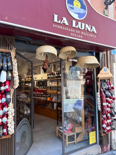 The image shows the exterior of a shop named "LA LUNA SOBRASSADA DE MALLORCA SOLLER" located in Palma de Mallorca, Spain. The shop specializes in cured meats, specifically sobrassada, which is evident from the display of numerous sausages hanging outside and visible through the window. The storefront features a maroon awning with the shop's name in large white lettering. Three woven straw baskets are suspended from the awning. Garlic braids are displayed on the left side of the entrance, and various packaged meats are visible on the right. The shop's interior displays shelves stocked with products, including bottles of wine and jars. A person is partially visible inside the shop. The street outside appears to be paved with tiles. A "Glovo" delivery service sticker is visible on the window.