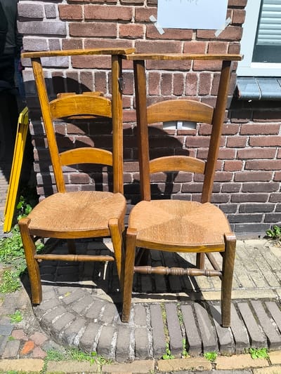 This image captures an outdoor scene in Alkmaar, Netherlands, featuring two wooden chairs positioned against a brick wall under daylight conditions.
Two high-backed wooden chairs with woven rush seats stand side-by-side. The chair on the left displays a lighter, more yellowish-brown wooden frame, while the chair on the right features a darker, richer brown wood tone. Both chairs have curved top rails and a curved lumbar support piece. Their seats are constructed from tightly woven rush with a distinct diagonal pattern. The chairs incorporate turned stretchers connecting their legs; the front stretcher of the right chair exhibits carved ring details. A metal hook is attached to the right vertical support of the left chair's backrest. The chairs cast strong, defined shadows onto the brick wall and the paved ground, indicating a light source from the upper left.
The background consists of a multi-toned brick wall, primarily composed of reddish-brown and darker grey-brown bricks laid in a stretcher bond pattern with visible mortar lines. A rectangular piece of white paper, bearing faint, illegible handwritten text, is taped to the upper section of the wall above the right chair, secured by two pieces of light-colored adhesive tape at its top corners. In the upper right corner of the frame, a segment of a white window frame with internal horizontal slats (likely blinds) and a dark, sloped sill is visible.
The ground is paved with rectangular bricks, exhibiting varying arrangements including longitudinal and arc patterns. Green weeds and small plants grow within the interstices of the bricks and along the edges of the paved surface. In the far-left periphery of the image, a bright yellow, multi-segmented object, possibly a folding ladder, is partially discernible. An obscured, dark shape, consistent with fabric or a partial human figure, is faintly visible in the extreme upper-left corner. No explicit human figures or active interactions are central to the scene.