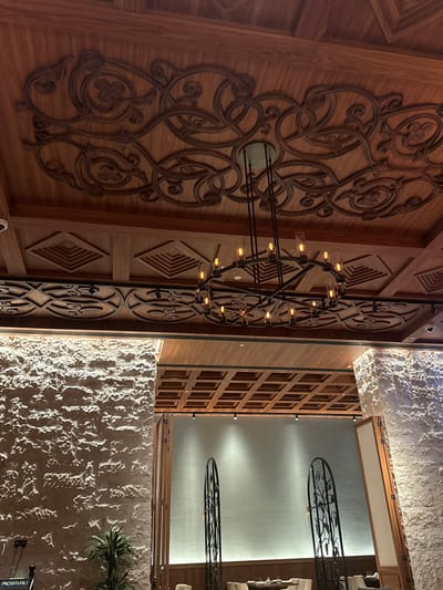 The image shows the interior of a building, likely in Doha, Qatar. A large, ornate wooden wall with carved designs dominates the left side of the image. A decorative metal chandelier with multiple small lights hangs near the center. To the right of the wooden wall is a section of textured white stone wall. Beyond the stone wall, a glimpse of the building's interior reveals wooden panels with small rectangular openings, possibly part of a decorative screen. Overhead, a glimpse of the building's ceiling reveals more decorative metalwork in an organic form. A plant is visible in the bottom right corner. The overall style suggests a luxurious, possibly Middle Eastern-inspired interior.
