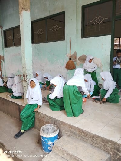 The scene depicts a covered outdoor area adjacent to a building in Jagapura Kulon, Indonesia. Ten individuals, consisting of nine young females and one young male, are present. The females are uniformly dressed in white long-sleeved shirts, green long skirts, and white headscarves. The male wears a white short-sleeved shirt and green long pants. The background wall is painted a distressed light green, exhibiting significant peeling; it features several dark-framed windows with decorative metal grates. A concrete support pillar is visible. A straw broom with a wooden handle leans against the wall. The floor is covered with light-colored square tiles. Most females are seated or crouching on the tiled floor or a concrete ledge, handling individual rectangular metal trays, small plastic containers with colored lids, and other items. One female in the left foreground sits on the concrete ledge, looking directly forward. In the right background, the male stands holding a stack of four small, shiny metal bowls. A blue and white plastic bucket, splattered with white paint, sits on the tiled floor near the concrete ledge.