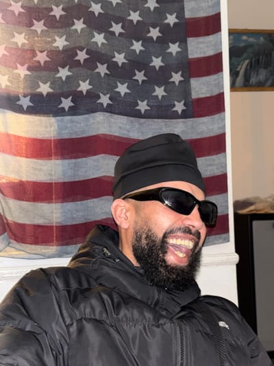 A man with a beard, wearing sunglasses and a black beanie, is laughing while standing in front of an American flag. He is also wearing a black puffer jacket. The location is unknown and cannot be determined from the image.