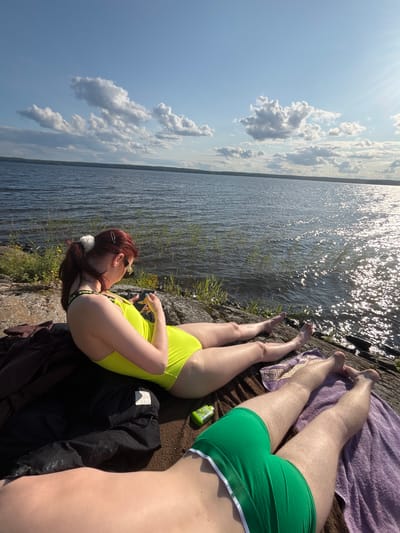 A full shot shows two individuals relaxing by a body of water in Shuiskaya Chupa, Russia, on a sunny day. In the foreground, a person wearing green swim shorts lies on a purple towel, facing away from the camera. Behind them, a woman in a neon yellow swimsuit reclines on a brown towel, looking down at her phone. She has her hair pulled back in a ponytail with a white scrunchie and is wearing sunglasses. A dark cloth is laid out on the rocks next to her. The background features a calm, dark lake with ripples and reflections of the sky. The sky is a bright blue with scattered white clouds, and the sun is visible, casting a warm glow on the water. The scene is set on a rocky shoreline with sparse green vegetation.