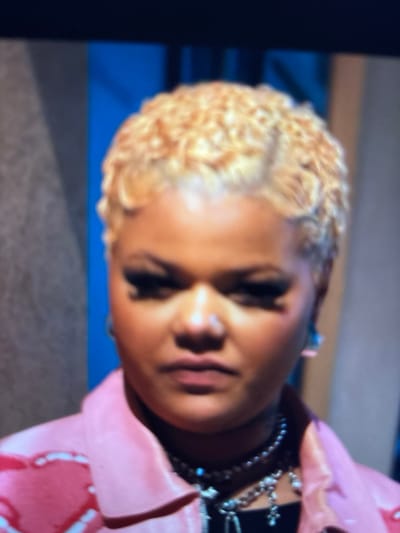 The scene is a close-up portrait of an adult female in Maisons-Alfort, France. She has a medium-dark complexion and short, light blonde, coiffed wavy hair. Her eyes feature prominent dark eyeliner and long false eyelashes. A small stud nose piercing is visible on her left nostril. She is wearing a shiny light pink jacket, potentially leather, with a distinct outlined or raised pattern on the left chest area, over a black top. Multiple silver-toned necklaces adorn her neck, including a pearl or beaded choker and longer chains with various pendants, one of which appears to be a small cross. A translucent, light-colored geometric earring hangs from her right ear. The blurred background consists of a vertical light grey textured panel on the left and a vertical bright blue section on the right, indicating an indoor, likely studio, environment.