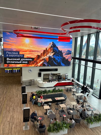 The image depicts the interior of an airport with a large digital screen displaying an advertisement for "Advertising at the airport: High level. Excellent view." The screen shows a mountain landscape with a sunset. Below the screen, a cafe area is visible with seating and tables. Several people are seated at tables in the cafe, some interacting with each other and others looking at their phones.

To the left of the cafe, a departure board is displayed, showing flight information. Further to the left, there are self-service kiosks. The floor is a light-colored wood laminate, and the ceiling features modern lighting fixtures and red decorative elements. Large windows on the right side of the image offer a view of the exterior, including trees and part of a parking lot.

The location is identified as the city of Старое Мартьяново, Russia, which is a small village in the Republic of Bashkortostan, Russia, and it is highly unlikely that this airport is located in such a small settlement. Therefore, the stated location is likely incorrect.