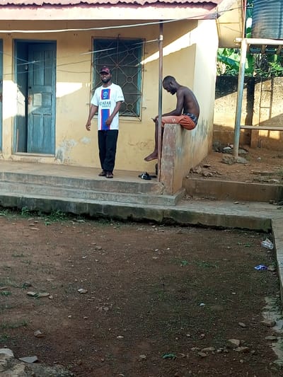 Two men are positioned outside a building. One stands, wearing a white and blue shirt, black pants, and a cap. He faces the camera. The second man sits shirtless on a low wall, looking down. The building has a yellow facade with a blue door and barred window. The ground is dirt with sparse grass and litter. A water tank is visible in the background.
