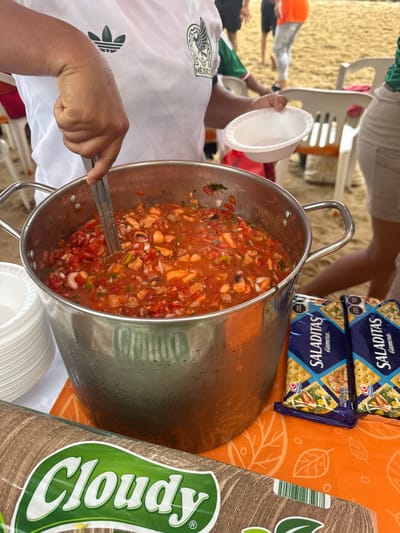 The image depicts a food service activity on a sandy beach in Acapulco, Mexico. A person, depicted from the mid-torso down, wears a white short-sleeved shirt adorned with an Adidas logo and the Mexico national football team crest on the left chest. This individual is actively serving a reddish-orange food mixture from a large, condensation-covered stainless steel pot. Their right hand manipulates a metal ladle within the pot, while their partially visible left hand holds an empty white disposable bowl, poised for receiving food. The pot's contents appear to be a mix of diced red tomatoes, white onion, green herbs, and various seafood pieces (both white and tentacled), consistent with a seafood cocktail or ceviche. To the left of the pot, a stack of multiple white disposable plastic bowls is present. The pot and bowls rest on a surface covered with an orange fabric patterned with lighter orange leaves. Two blue packages of "Saladitas Gamesa" crackers, featuring images of crackers with toppings, are positioned to the right of the pot. In the bottom-left corner, a partially visible product package labeled "Cloudy" with a green logo displays a wood-grain texture. The background shows blurred figures, one in an orange top, and several white plastic chairs scattered across the sandy beach.